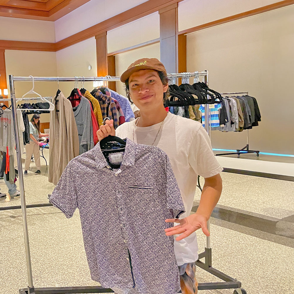 Student holding a short sleeve shirt on a hanger, with clothing racks behind him at a Swap Shop event