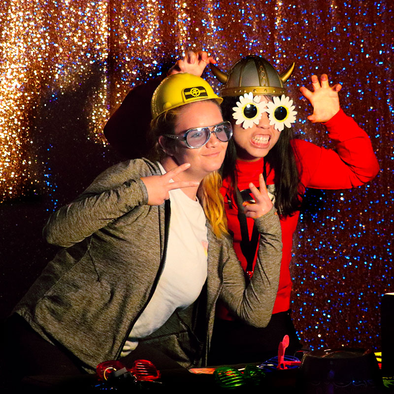 Two students wearing funny glasses and hats at a photo booth
