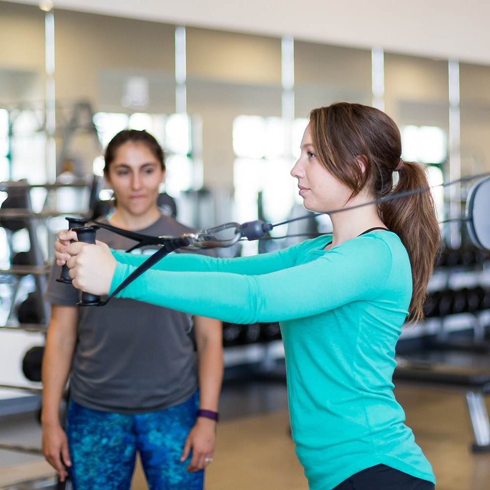 A woman using a weight machine to work out while a trainer spots her.
